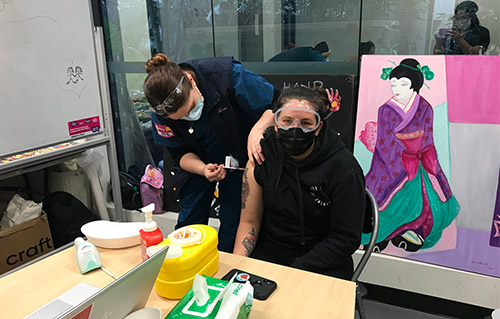 Women wearing masks, one is looking toward the camera and receiving the vaccine while the other is administering the vaccine. 
