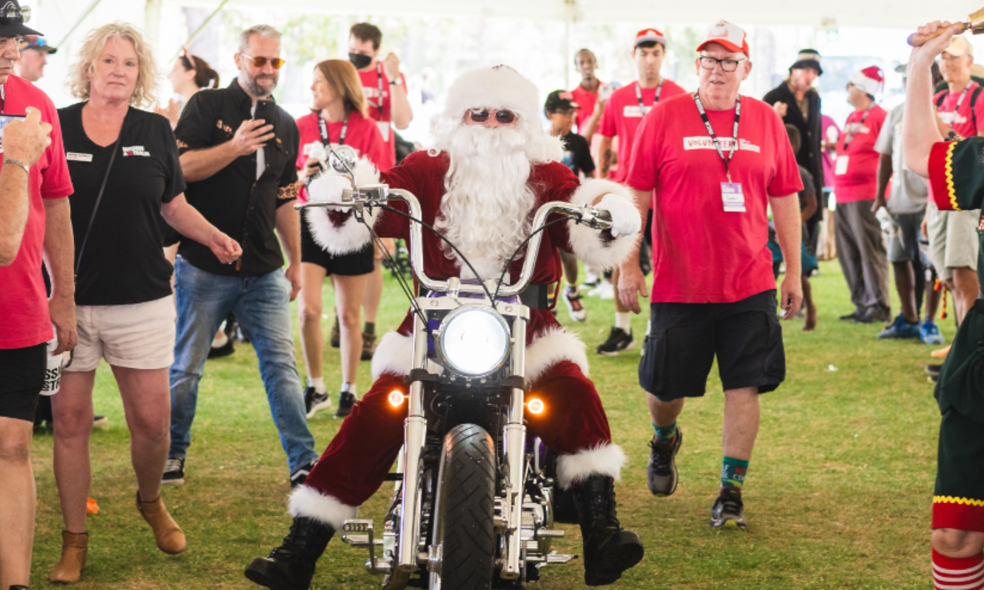 Santa Claus rides a motorcycle through a crowd at a Christmas event, surrounded by volunteers in red shirts.