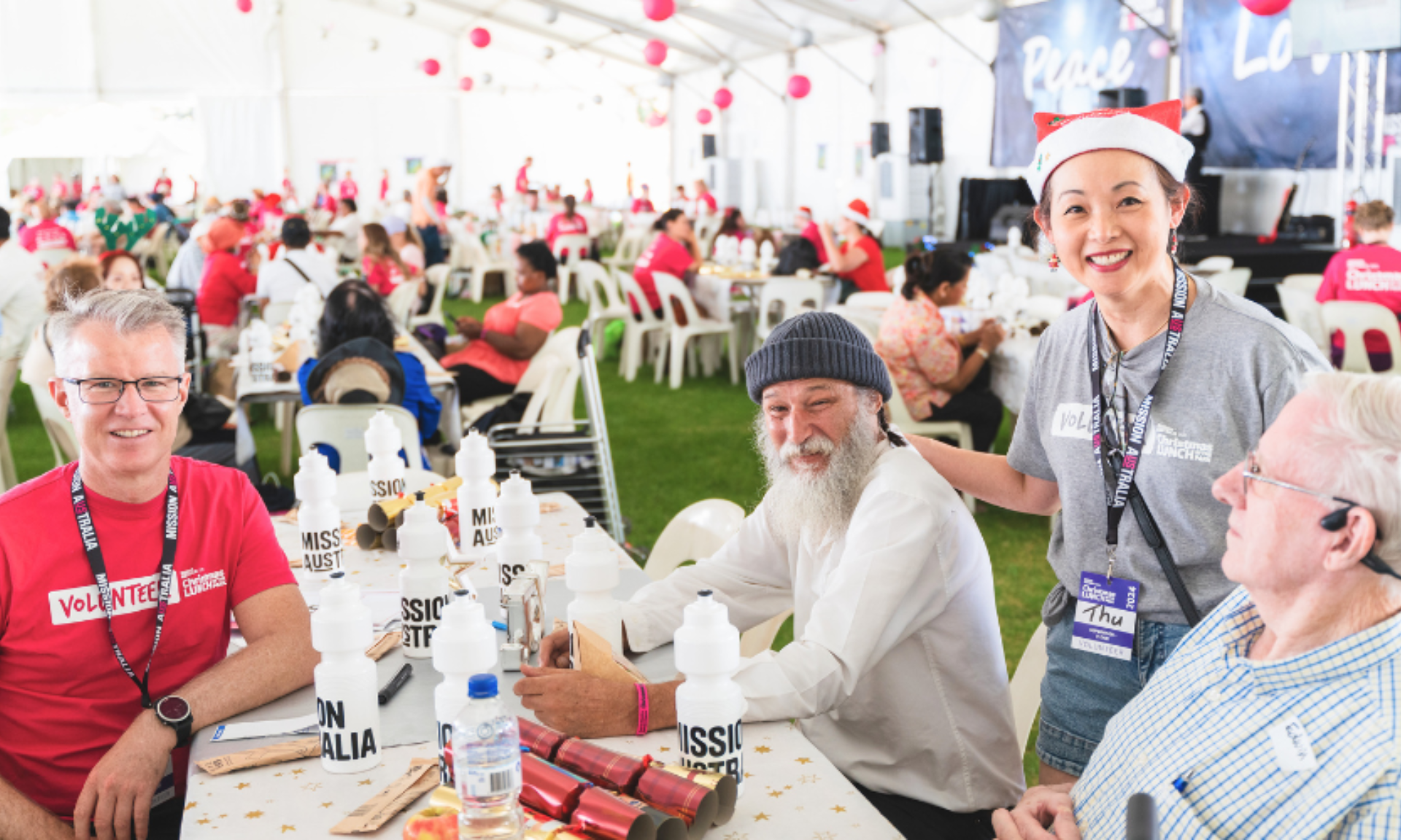 Volunteers and guests sit at a table with Mission Australia water bottles during a Christmas lunch event under a decorated tent.