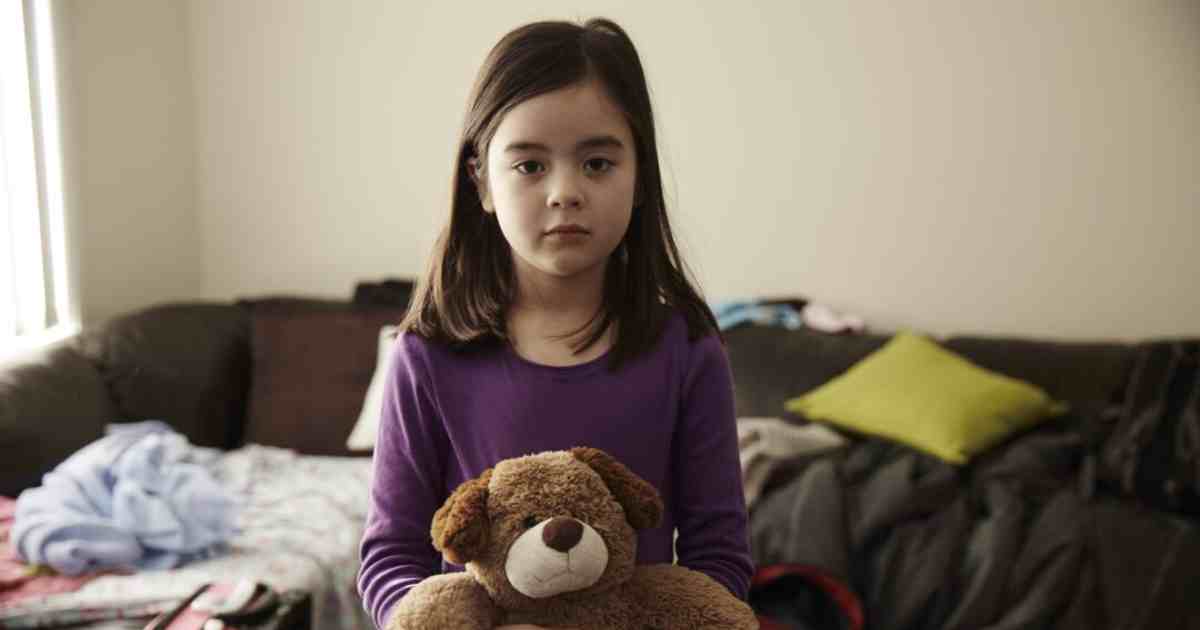 young child holds her teddy with her belongings in the background