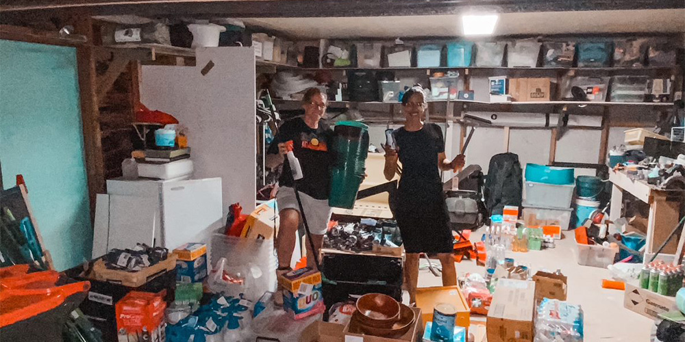 Two Mission Australia workers standing inside a garage filled with essential items such as food, cleaning products and fuel to donate families in the Coffs Harbour community.  