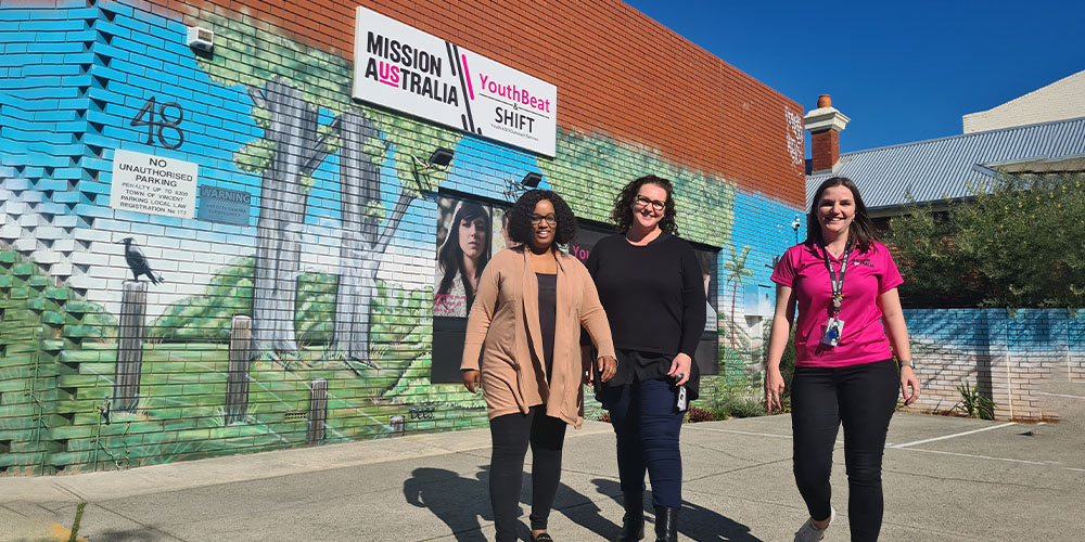 Three women smiling and walking in front of a Mission Australia building.