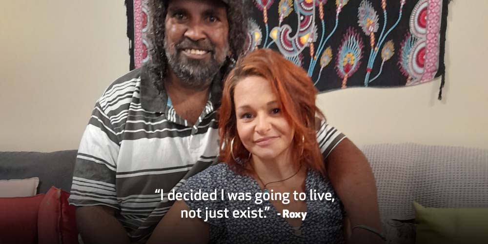 Roxy smiles at the camera with her partner. Text on image reads ’I decided I was going to live, not just exist.” 
