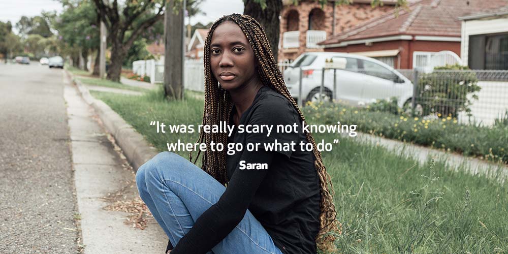 A worried Saran sits on the side of the road with her hands clasped around her knees.