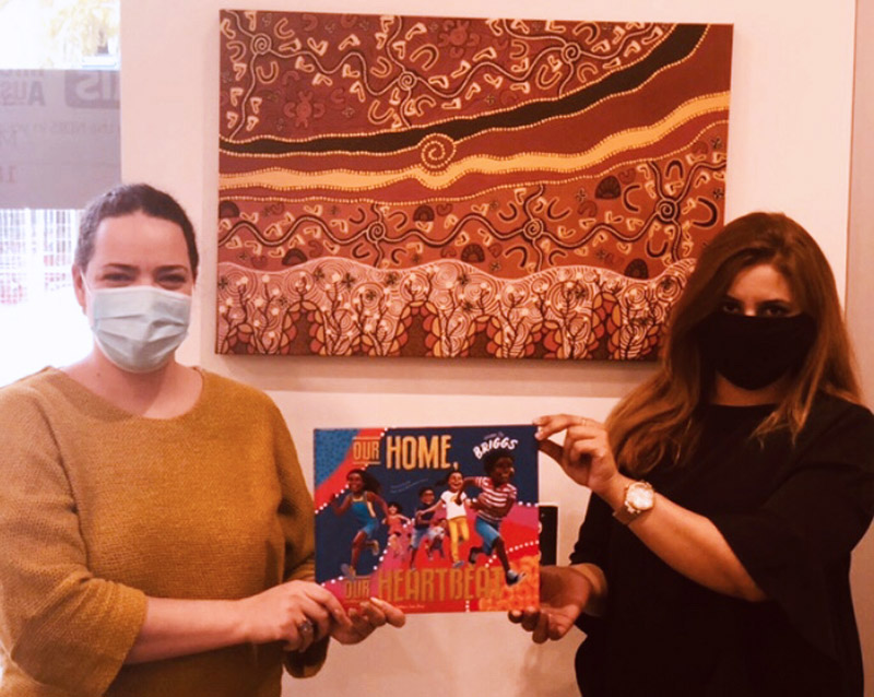 Women smiling at the camera, wearing masks and holding the book ’Our Home, Our Heartbeat’. 